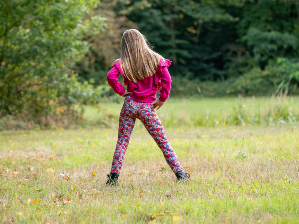 meisje met een handgemaakte roze sweater aan en een legging met vrolijke bloemetjes. je ziet haar van de achterkant