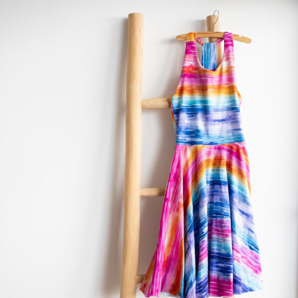 Halterjurk voor kinderen met pastel regenboogstreepjes en zwierige cirkelrok, hangend op houten ladder met witte achtergrond